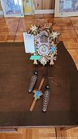 Full view of the wooden cuckoo clock showing clock face, carved bird, floral hand painting, and pinecone weights and pendulum on table.