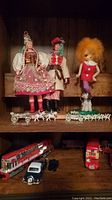 First image displaying dolls, miniature horse-drawn carriages, model train locomotive, toy cars, and a double-decker bus arranged on wooden shelves.