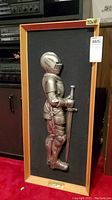 Framed knight sculpture in full armor holding a sword, shown upright within a wooden frame, on red carpet