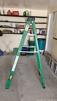 Green metal foldable step ladder fully open, showing frame and steps in garage environment.