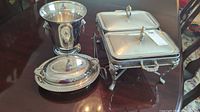 Photo showing a silver plated ice bucket, a covered round serving dish, and the double covered chafing dish on a wooden table