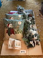 Two snowman-themed Christmas pillows, a holiday garland with pine cones and white berries, and two white hand towels with Christmas tree embroidery arranged on floor