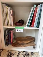 Cookbooks arranged vertically and horizontally on a shelf, wooden container with lid beside books, wooden serving bowl on lower shelf.