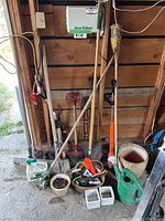 Garden tools including shovels, rake, hoe, pitchfork with wooden handles, garden pots, and green watering can against garage wall