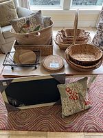 Photo of assorted woven baskets and decorative pillows with embroidered birds on a table along with round wooden trivets and a framed chalkboard on the floor.