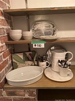 View of all items on shelves including white ceramic bowls, plates, metal serving trays, and mugs with utensils.