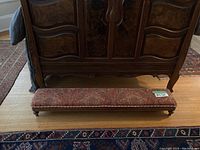 Long upholstered low rise foot stool positioned in front of a wooden dresser, showing full length and one side view.