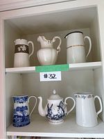 Front view showing all five pitchers and one teapot arranged on two shelves in a cabinet.