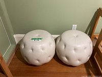 Front view of two white pleather ottoman poufs side by side on wood floor against green wall