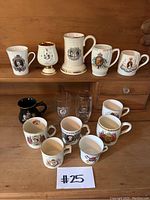 Full set of 14 commemorative Royal Family cups and mugs on wooden shelf, showing variety of designs and colors