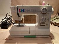 Front view of white Janome sewing machine showing stitch selection chart, dials for stitch length and reverse, threaded needle, power cord attached, placed on beige surface.