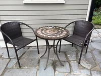 Full set showing two brown wicker chairs and one round table with a stone mosaic top and black metal frame on outdoor stone patio.