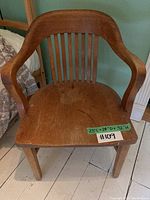 Front view of vintage wooden desk chair showing seat, slatted backrest, and armrests with a green label for dimensions and lot number.