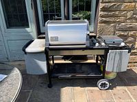 Front view of Weber Genesis Gold propane barbecue grill showing silver lid, white side burner, and side table with wheels and propane tank underneath.
