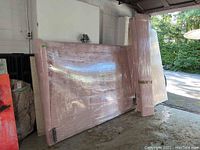 Wide shot of shrink-wrapped pink upholstered headboard and side rails against garage wall