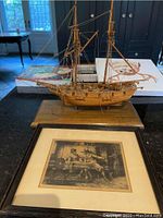 Side angle view of wooden ship model and framed engraving print together on a table.