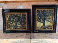 Two framed oil paintings side by side showing dark tree scenes, black and gold frames, set against a light wall.