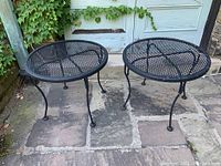 Two round black metal patio side tables with mesh tops positioned side by side on stone patio in front of a door with ivy on the wall.