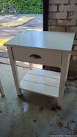 Front and side angled view showing the white painted wood side table with a drawer and lower shelf.
