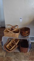 Seven woven baskets arranged on floor against wall including large rectangular basket with handles, three round basket bowls, small divided basket, dark brown basket, and two more baskets.