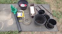 Photo showing green Barracuda blower, yellow portable light, black bucket with items, metal trivet, wooden trays, and multiple black plastic pots and saucers on a mat outdoors.