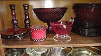 Wide shot of cranberry glass items on shelves including candlesticks, bowls, plates, jar candle, and gravy boat.