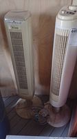 Two standing fans, one Hawaiian Breeze tower fan about 48 inches tall and one smaller table fan, both dusty and needing cleaning