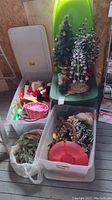 Three large plastic storage bins and a white plastic bag containing various holiday decorations such as artificial Christmas trees, wreaths, decorative berry stems, and Santa figurines.