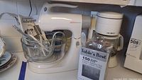 Sunbeam Mixmaster mixer with glass bowl, user manual, Mr. Coffee coffee maker, and boxed cutlery set on table.