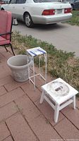 Photo showing white wooden stool, white metal plant stand with tile top, and large planter pot beside a patio area.