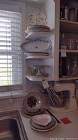 Stacked plates and serving dishes on kitchen shelves by window with light coming through. Several pieces visible with floral and gold patterns.