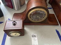 Photo showing both clocks on a white surface. The wooden electric mantel clock has a curved silhouette and the brown leatherette battery table clock has a small handle on top.