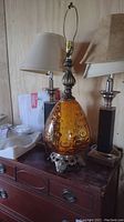 Photo showing the amber glass MCM lamp base in front of two 30-inch leather-like and silver base table lamps with white shades on a wooden dresser.