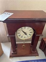 Full frontal view of the wooden tabletop pendulum clock showing the classic rectangular body, supporting columns, white clock face with Roman numerals, and brass pendulum beneath.