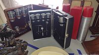 Photo showing two portable bar setups on a table. On the left is a wooden or metal case with six stacked metal cups and bar accessories such as corkscrew and stirring spoon mounted inside black foam. On the right, two silver metal Thermoses with red lids and plastic figures are visible with a red travel bag.