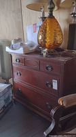 Front view of vintage wooden three drawer buffet with lamp on top, a few other items and a chair beside it.