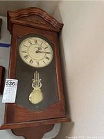 Front view of wooden pendulum wall clock showing clock face with Roman numerals and visible pendulum inside glass door.