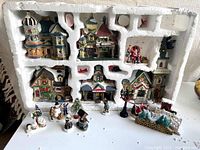 Overview photo displaying six Christmas village buildings in the original styrofoam packaging accompanied by assorted smaller figures and decorations arranged on a white surface.