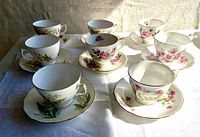Eight English porcelain teacup and saucer sets arranged on a white cloth, showing floral patterns and gold trim. No chips or cracks visible.