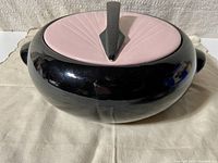 Front angled view of the pink lid with sun dial design and black body casserole dish on cloth background.