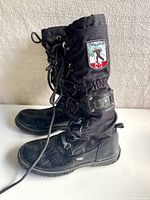 Side profile of black tall winter boot showing leather bottom, textile upper, and lace-up front with Pajar logo patch.