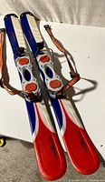 Top view of the two snow blades showing the red, white, and blue color pattern on the surface and the adjustable orange straps with bindings.