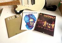 Three songbooks displayed next to a white electric guitar showcasing covers for Elvis Presley, David Bowie, and The Who
