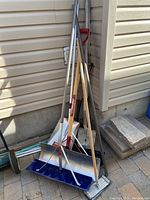 Photo showing various garden tools including shovels, rakes, a large metal push shovel with blue edge, and a wall sander leaning against house siding on outdoor ground with bricks and stone slabs nearby.
