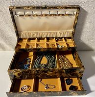 Vintage jewelry box open showing top pull-out compartment with small sections containing several pairs of earrings. The lid has necklace hooks and butter velvet lining.