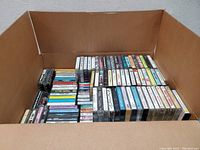 Photo of large box filled with assorted cassette tapes organized in stacks, visible titles include various musical genres and artists.
