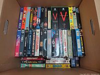 Photo showing a box containing 31 VHS tapes arranged upright with titles visible including V, Videodrome, Spacehunter, Cherry 2000, The Fantastic Planet, and others.