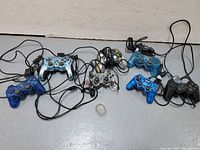 Seven wired PS2 controllers laid out on a floor; various colors and designs visible including one with palm cooling fan.