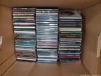 Photo showing a large box filled with stacks of music CDs in jewel cases, arranged in multiple vertical stacks, mostly spine-up to show artist/title information.