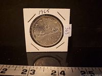 Front side of 1965 Canadian silver dollar showing the canoe design with two voyageurs and the year 1965 in a white protective holder.
